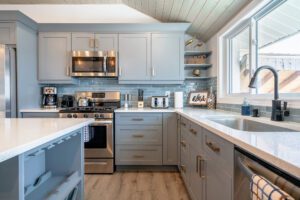 This is an example of Dream BNB Hospitality luxury rental property with a beautifully designed kitchen with grey cupboards and clean white countertops; farm house sink with view overlooking the Lake Erie waterfront; stainless finishes on fridge, stove, microwave, dishwasher and coffee pot; shiplap ceiling and light colored wood flooring. 