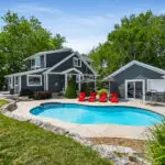 This is an example of Dream BNB Hospitality luxury rental property with essential vacation rental amenities, including a beautifully designed luxury lakefront cottage on Lake Erie, Ontario; two-story waterfront home with navy blue siding and white trim; inground saltwater pool; red sun loungers; neat and modern landscaping with tall grasses and rock borders.