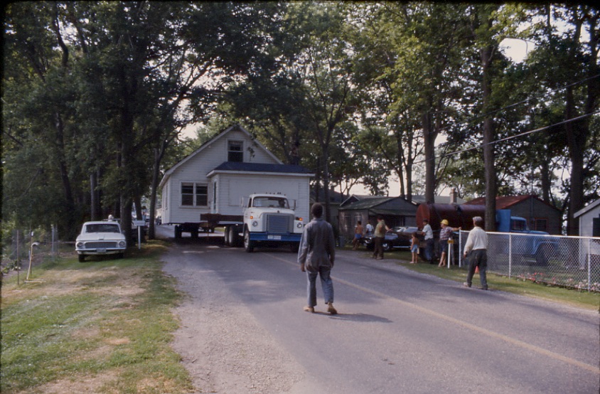 View on Point Pelee Drive of a small cottage on a flat-bed truck being relocated from Point Pelee National Park in the 1960s.